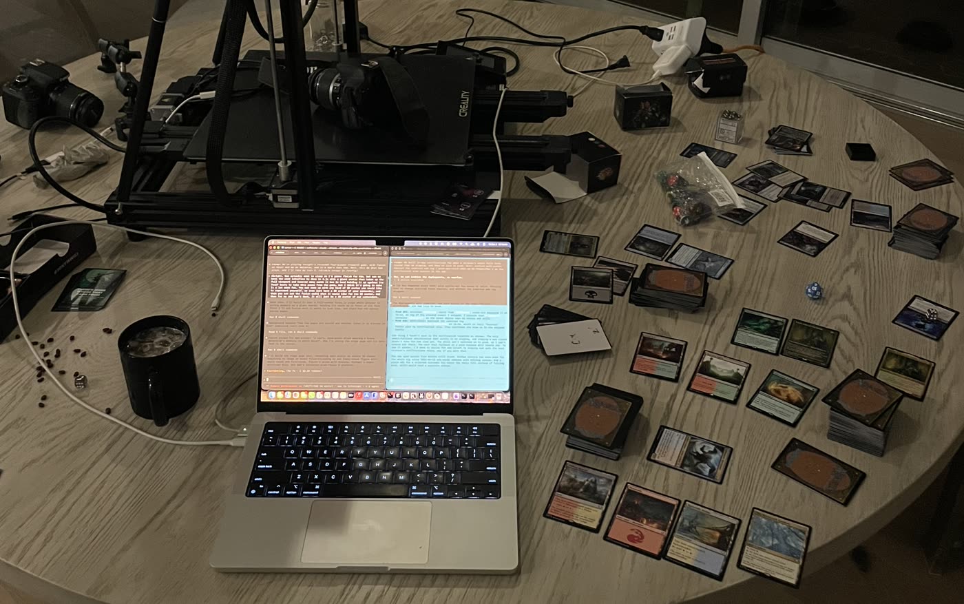 A round wooden table at night: a laptop running the table, a 3D-printer camera gantry, decks, dice and Magic cards in play.